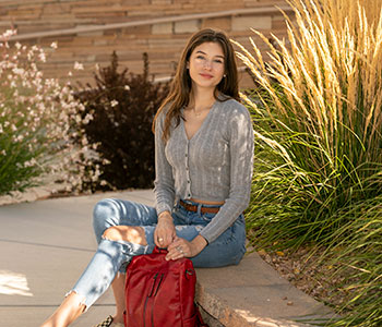 Student holding red backpack and sitting on wall surrounded by native landscape