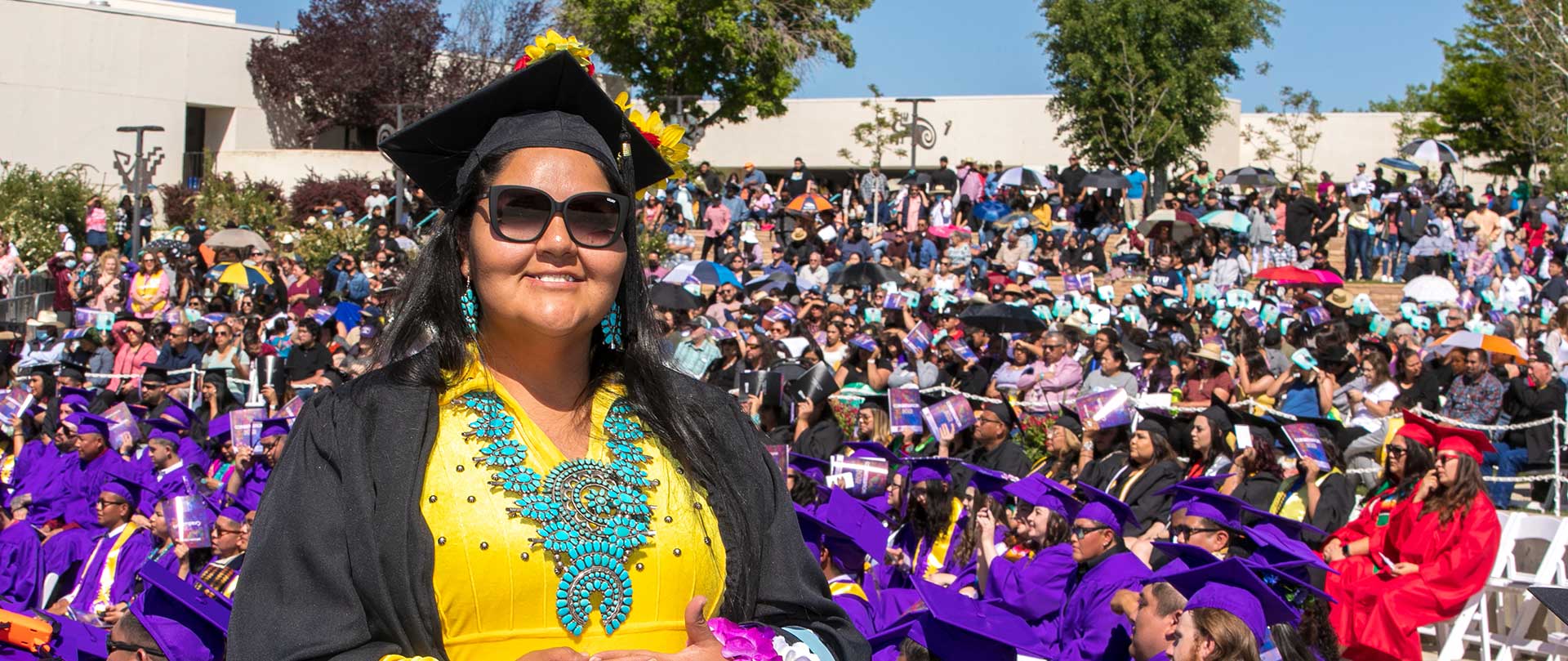 SJC Graduate wearing cap, sunglasses, and  native jewelry smiling at camera with graduates in background